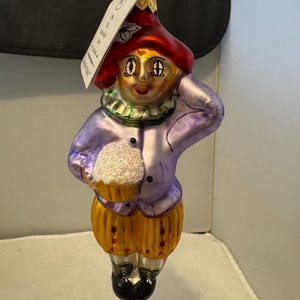 Puede incluir: Adorno de cristal caprichoso con forma de personaje sosteniendo una magdalena. El adorno presenta un sombrero rojo, una chaqueta morada y pantalones amarillos. La magdalena tiene una cobertura blanca y brillante. Zapatos negros completan el look.