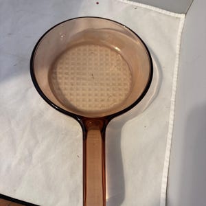 May include: An amber-coloured glass saucepan with a single handle. The pan has a round shape with a textured base. The handle is a solid piece of glass, and the pan is designed for cooking.