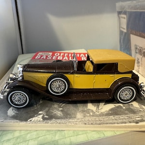 May include: A brown and tan vintage car model with a tan fabric roof. The car is sitting on a book with a red and white label that says "Official Baseball Rules".
