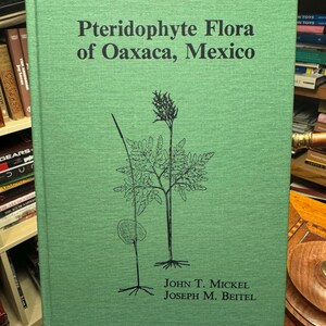 Peut inclure: Un livre relié avec une couverture texturée vert clair. Le titre Pteridophyte Flora of Oaxaca, Mexico est imprimé en noir. Sous le titre se trouve un dessin au trait noir représentant deux plantes. Les auteurs John T. Mickel et Joseph M. Beitel sont indiqués en bas en noir.