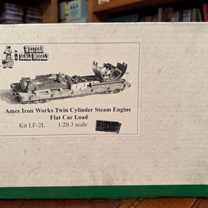 Pode incluir: Uma caixa para um kit Western Scale Models. A caixa apresenta uma ilustração a preto e branco de um motor a vapor de dois cilindros Ames Iron Works Flat Car Load. O texto na caixa diz "Kit LF-2L" e "escala 1:20.3".