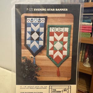 Peut inclure: Un emballage présentant deux patrons de bannières patchwork. L'une est bleue et blanche avec des cœurs et des tulipes, l'autre est rouge, verte et blanche avec le mot "NOEL". L'emballage indique "P-121 EVENING STAR BANNER". La bannière mesure 35,56 cm x 66,04 cm.