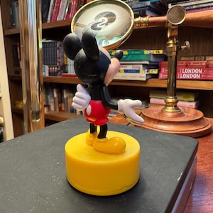 Puede incluir: Una figura de Mickey Mouse sobre una base amarilla. La figura tiene una cabeza negra con orejas grandes, guantes blancos, pantalones cortos rojos y zapatos amarillos. El fondo incluye una lupa y estanter&iacute;as.