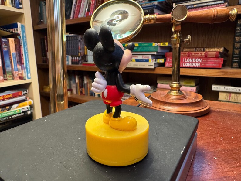 Puede incluir: Una figura de Mickey Mouse sobre una base amarilla. La figura tiene una cabeza negra con orejas grandes, guantes blancos, pantalones cortos rojos y zapatos amarillos. El fondo incluye una lupa y estanter&iacute;as.