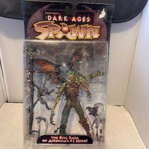 Peut inclure: Figurine Dark Ages Spawn dans son emballage d'origine. La figurine est une représentation détaillée et multicolore d'une créature ailée avec un bâton et d'autres accessoires. L'emballage indique "The Epic Saga of America's #1 Hero!"