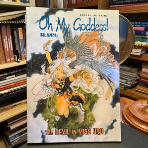 May include: A manga book titled Oh My Goddess! The Devil in Miss Urd by Kosuke Fujishima. The cover features a central illustration of a character with long white hair, wearing orange and black clothing, accompanied by a large bird with spread wings.