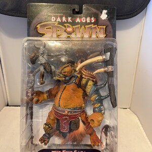 Puede incluir: Figura de acción de Dark Ages Spawn en un paquete de plástico transparente. La figura es una criatura grande, de color marrón anaranjado, con cuernos, armadura y un arma. El embalaje tiene las palabras "Dark Ages Spawn" y "The Epic Saga of America's #1 Hero!"