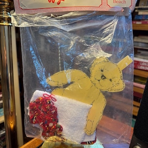 May include: A yellow teddy bear cutout holding a white towel with a red and multicolored trim, packaged in a clear plastic bag. The packaging reads "The Muffy Collection" and "At The Beach".