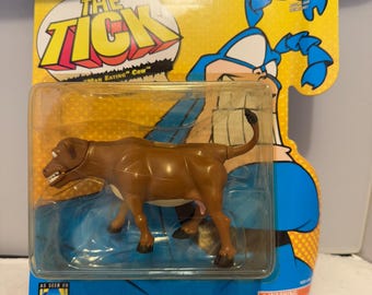 May include: A brown action figure of a cow from "The Tick" cartoon, packaged on a yellow and blue card. The packaging features the show's logo and the Fox Kids Network logo. The figure is in a clear plastic case.