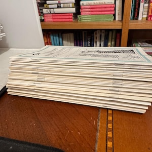 Op de afbeelding: Een stapel vintage nieuwsbrieven met een blauwe rand en zwart-wit illustraties. De nieuwsbrieven liggen op een donkere houten tafel, met een boekenkast vol boeken op de achtergrond.