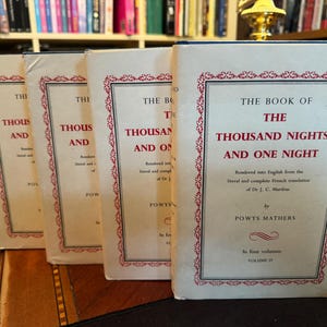 May include: Four vintage books titled "The Book of the Thousand Nights and One Night" are displayed. The book covers are cream-colored with red and black text and decorative borders. The books are arranged on a wooden surface.