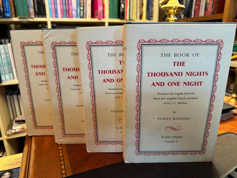 May include: Four vintage books titled "The Book of the Thousand Nights and One Night" are displayed. The book covers are cream-colored with red and black text and decorative borders. The books are arranged on a wooden surface.