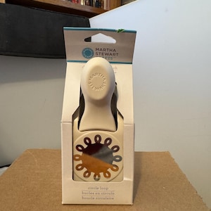 May include: A Martha Stewart circle loop paper punch in its original packaging. The punch is off-white with a decorative circle loop design in shades of brown, orange, and blue. The packaging includes the text "circle loop, bucles en círculo, boucle circulaire."