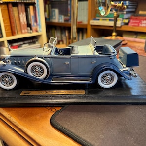 May include: A blue and grey 1931 Cadillac Sport Phaeton model car featuring white-walled tires, wire-spoke wheels, a spare tire mounted on the side, and a trunk attached to the rear. The vehicle is displayed on a rectangular black wooden base.