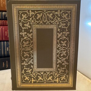 Könnte beinhalten: Ein dunkelbraunes Hardcover-Buch mit goldenen Verzierungen. Der Einband hat einen goldenen Rahmen mit floralen und geometrischen Mustern. Das Buch ist Teil einer Sammlung antiker Bücher.