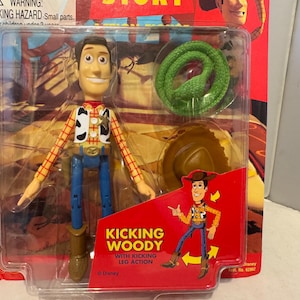 May include: A Woody action figure from the movie Toy Story, packaged in a clear plastic case. The figure is wearing a cowboy hat, vest, and blue jeans. The packaging includes a green snake and a hat. The text "Kicking Woody" is visible.