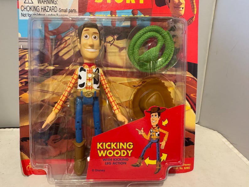 May include: A Woody action figure from the movie Toy Story, packaged in a clear plastic case. The figure is wearing a cowboy hat, vest, and blue jeans. The packaging includes a green snake and a hat. The text "Kicking Woody" is visible.