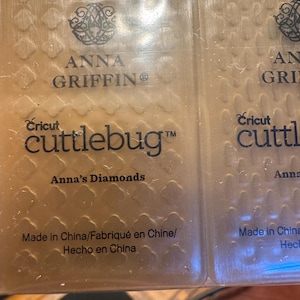 Może przedstawiać: Dwie przezroczyste plastikowe teczki do wytłaczania Cricut Cuttlebug. Teczki mają logo Anny Griffin i tekst "Anna's Diamonds". Widoczny jest również tekst "Made in China/Fabriqué en Chine/Hecho en China".