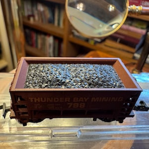 May include: A miniature brown model train coal car filled with black coal. The words "THUNDER BAY MINING 798" are printed on the side of the car. The car is sitting on a clear surface.