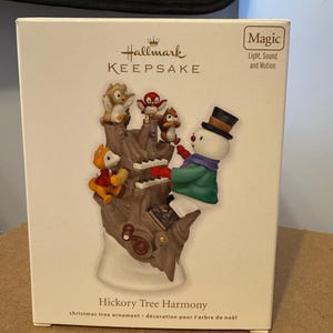 May include: Hallmark Keepsake Christmas ornament featuring a snowman playing a piano on a tree stump with a squirrel, owl, and fox. The ornament is titled "Hickory Tree Harmony" and features light, sound, and motion.