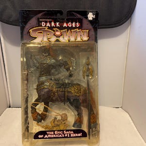 以下が含まれることがあります： オリジナルパッケージに入ったSpawn Dark Agesのアクションフィギュア。このフィギュアは、鎧と槍を持ったケンタウロスのようなキャラクターを描いています。パッケージには「Spawn Dark Ages」と「The Epic Saga of America's #1 Hero!」と書かれています。