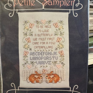 Può includere: Un campionario di punto croce con sfondo bianco e accenti floreali rosa e verdi. Il motivo include il testo "Per poter amare una farfalla, dobbiamo prima prenderci cura di alcuni bruchi." e l'alfabeto. Due coniglietti marroni sono cuciti nella parte inferiore del motivo.