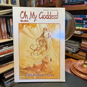 May include: A softcover manga book titled Oh My Goddess! Childhood's End by Kosuke Fujishima. The white cover features a warm-toned illustration of three female characters with long flowing hair in a layered composition with golden yellow hues.