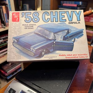 Könnte beinhalten: Ein AMT '58 Chevy Impala Modellbausatz-Karton mit einer blauen Auto-Illustration und dem Text '58 CHEVY IMPALA BUILD STOCK, CUSTOM OR DRAG'. Der Karton steht auf einer digitalen Accuteck-Waage, die 0,105 Pfund anzeigt.