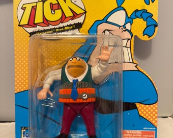 May include: A packaged action figure of "The Tick" character, featuring a Dyna-Mole design. The figure is in a clear plastic case, with a yellow and blue backing card. The figure is wearing a green vest, red pants, and a blue tie.