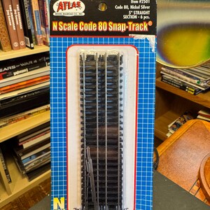May include: Atlas Model Railroad Co. N Scale Code 80 Snap-Track, item #2501. This package contains 6 pieces of 5-inch straight nickel silver track sections. The product is mounted on a blue grid-patterned cardboard backing with white text.