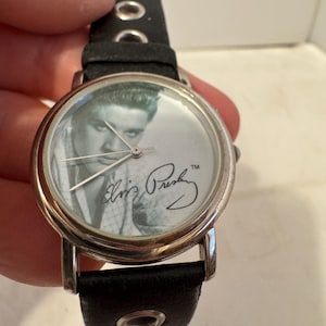 May include: A black leather wristwatch with a silver face featuring a black and white photo of Elvis Presley and his signature. The watch has a silver band with a black leather strap.