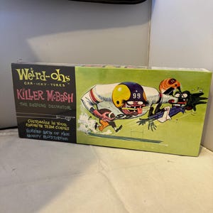 Pode incluir: Uma caixa vintage para um kit de modelo "Weird-ohs" com um jogador de futebol de desenho animado com o texto "Killer Mobash, The Dazzling Decimator". A caixa é verde e amarela com uma ilustração de desenho animado de um jogador de futebol correndo com a bola. A caixa também inclui o texto "Customize in your favorite team colors, Horrible bits of fine mouldy polystyrene."