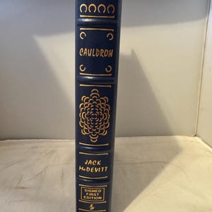Può includere: Un libro rilegato in blu con lettere dorate. Il titolo è "Cauldron" di Jack McDevitt. Il libro è una prima edizione firmata.