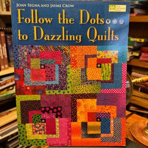 Op de afbeelding: Een boek getiteld "Follow the Dots to Dazzling Quilts" van Joan Segna en Jayme Crow. De omslag toont een kleurrijk patchwork-ontwerp met vierkanten in tinten rood, groen, paars en oranje. Het boek ligt op een donkere ondergrond.