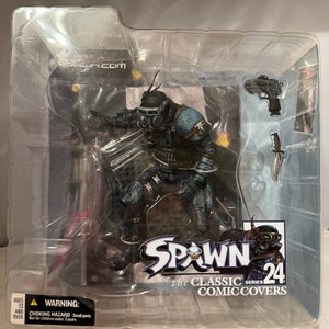 May include: A Spawn action figure from the Classic Comic Covers series, number 24, in its original packaging. The figure is a detailed, dark-colored character with a helmet, mask, and armor. Accessories include a gun and a knife. The packaging includes a choking hazard warning.