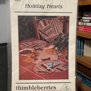 May include: A sewing pattern for holiday-themed heart-shaped quilted coasters. The pattern is called "Holiday Hearts" and is from the company "thimbleberries". The pattern shows a red and white checkered tablecloth with three heart-shaped coasters and a larger heart-shaped pillow.