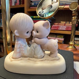 Może przedstawiać: Ceramiczna figurka przedstawiająca dziecko i świnkę w uścisku. Dziecko ma na sobie jasnoniebieski i różowy strój, świnka jest jasnoróżowa. Podstawa jest w kolorze złamanej bieli. Temat jest sentymentalny.