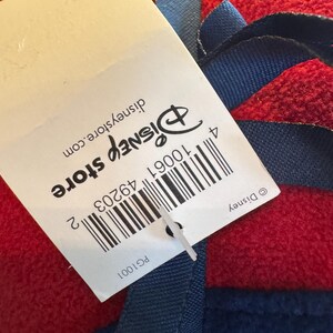 K&ouml;nnte beinhalten: Eine rote Decke mit blauen Zierleisten und einem blauen Band. Ein wei&szlig;es Etikett mit dem Disney Store-Logo und Barcode ist an der Decke befestigt. Das Etikett enth&auml;lt auch den Text "disneystore.com".