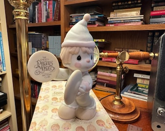 May include: A Precious Moments figurine of a child in a white outfit and hat, holding a round sign that reads "The Enesco Precious Moments Birthday Club". The figurine has a light pink trim and is standing on a patterned surface.