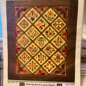 May include: A quilt pattern titled "Hugs and Kisses From Mackenzie" with a patchwork design featuring a variety of floral and geometric motifs in shades of red, orange, yellow, green, and brown. The pattern is 64 inches by 75 inches and includes the text "From my heart to your hands" and "Quilt Designs by Lori Smith".