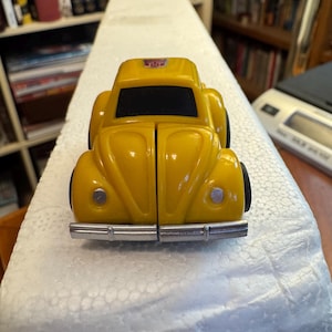 Könnte beinhalten: Ein gelbes Spielzeugauto mit abgerundeter Karosserie, schwarzen Fenstern und einer silbernen vorderen Stoßstange. Das Fahrzeug hat ein kleines violett-silbernes Emblem auf dem Dach und runde silberne Scheinwerfer an den vorderen Kotflügeln.