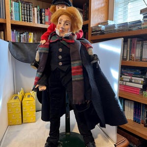 Puede incluir: Una muñeca detallada de un hombre con abrigo, chaleco y pantalones negros, con una bufanda estampada. Una muñeca más pequeña está sentada sobre sus hombros, con una chaqueta roja y un sombrero negro. Las muñecas están sobre un soporte, con estanterías de fondo.