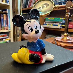 Pu&ograve; includere: Statuetta in ceramica di Topolino seduto. La figura ha orecchie nere, viso bianco e camicia blu. Sono visibili anche i pantaloncini rossi e le scarpe gialle di Topolino. La statuetta &egrave; un oggetto da collezione.