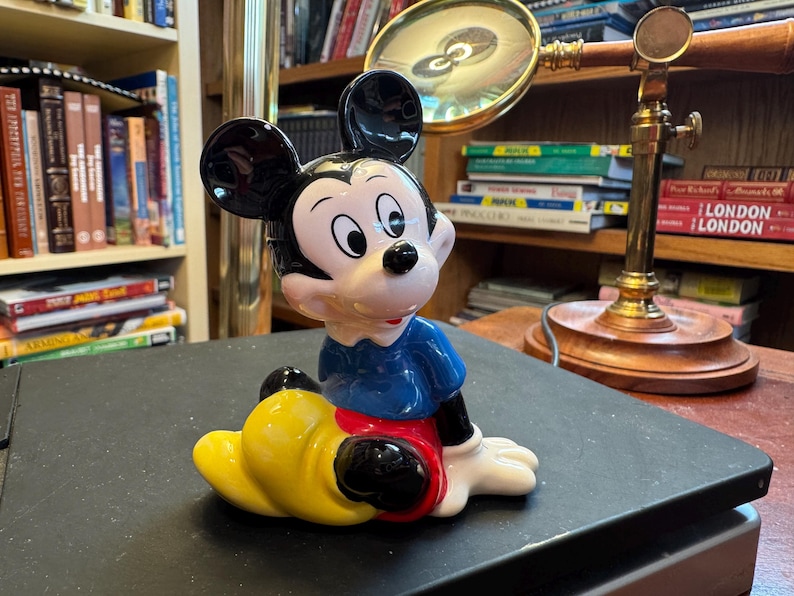 Pu&ograve; includere: Statuetta in ceramica di Topolino seduto. La figura ha orecchie nere, viso bianco e camicia blu. Sono visibili anche i pantaloncini rossi e le scarpe gialle di Topolino. La statuetta &egrave; un oggetto da collezione.