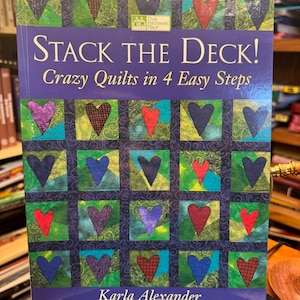 May include: A book titled "Stack the Deck! Crazy Quilts in 4 Easy Steps" by Karla Alexander. The cover features a grid of colorful heart-shaped quilt blocks in various colors, including red, purple, and blue, set against a green and blue background.