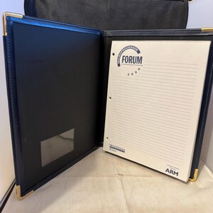 K&ouml;nnte beinhalten: Ein blauer Lederordner mit goldener Einfassung und einem wei&szlig;en Notizblock im Inneren. Der Notizblock hat einen blauen Kreis mit dem Text "MICROPROCESSOR FORUM 2000" und ein Logo f&uuml;r MicroDesign.