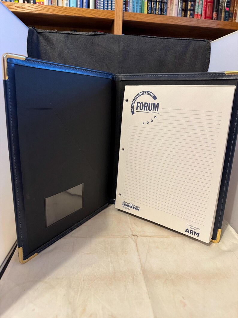 K&ouml;nnte beinhalten: Ein blauer Lederordner mit goldener Einfassung und einem wei&szlig;en Notizblock im Inneren. Der Notizblock hat einen blauen Kreis mit dem Text "MICROPROCESSOR FORUM 2000" und ein Logo f&uuml;r MicroDesign.