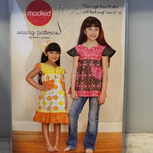 May include: A sewing pattern for a tunic-style dress and shirt with a head scarf. The pattern is called "Frida" and is from the "modkid sewing patterns" line by Patty Young. The pattern is for sizes 2T-10.