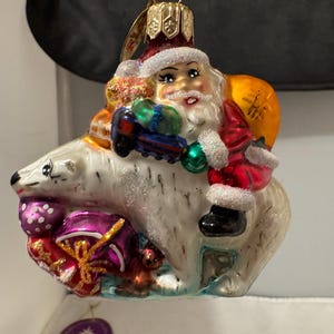 May include: A glass Christmas ornament featuring Santa Claus riding a polar bear. Santa is wearing a red suit with white trim and a white beard. He is holding a bag of gifts and a toy. The polar bear is white with a blue and green scarf. The ornament is decorated with glitter and snowflakes.