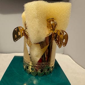 Könnte beinhalten: Ein goldfarbener Kerzenhalter mit Engelmotiv und zylindrischer Form. Der Halter hat dekorative Ausschnitte und Engelsflügel. Im Inneren des Halters befindet sich ein gelber Schwamm. Der Kerzenhalter steht auf einer türkisfarbenen Oberfläche.